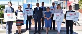 Na zdjęciu znajduje się 9 elegancko ubranych osób. Większość z nich trzyma w rękach symboliczne promesy ze środkami wsparcia finansowego z Rządowego Funduszu Inwestycji Lokalnych. 