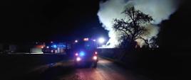 Zdjęcie przedstawia zastępy straży pożarnej podczas gaszenia pożaru stogu słomy. Na drodze stoją samochody strażackie z włączonymi światłami błyskowymi koloru niebieskiego. Za płotem betonowym widoczny dym, który unosi się do góry.