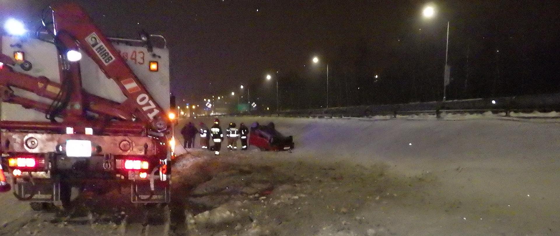Zdjęcie przedstawia samochód, który znajduje się na dachu. Na pierwszym planie po lewej stronie wóz strażacki.