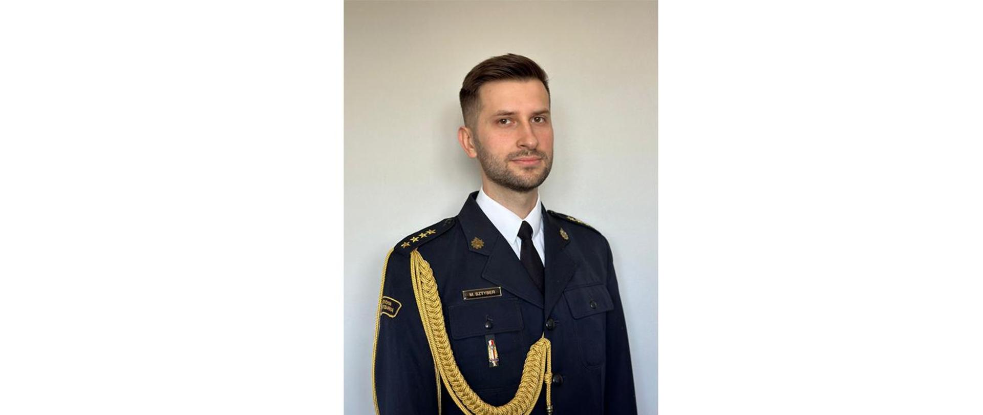 Zdjęcie portretowe p.o. Zastępcy Komendanta Powiatowego Państwowej Straży Pożarnej w Krośnie Odrzańskim (st. kpt. Michał Sztyber) na jasnym tle w mundurze galowym.