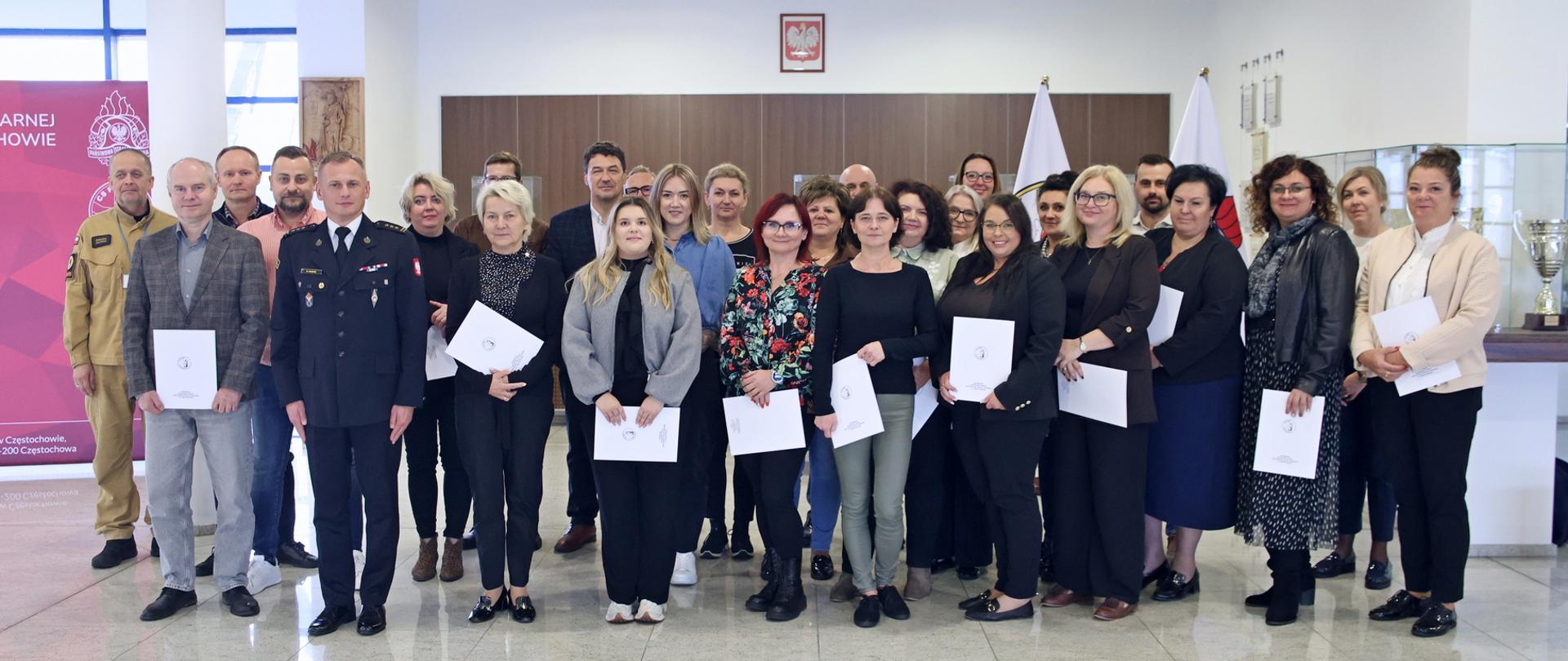 Pamiątkowe zdjęcie grupowe uczestników szkolenia z zakresu ochrony ludności i obrony cywilnej (25 osób) w towarzystwie Zastępcy Komendanta Centralnej Szkoły PSP, kobiety oraz funkcjonariusza CS PSP
