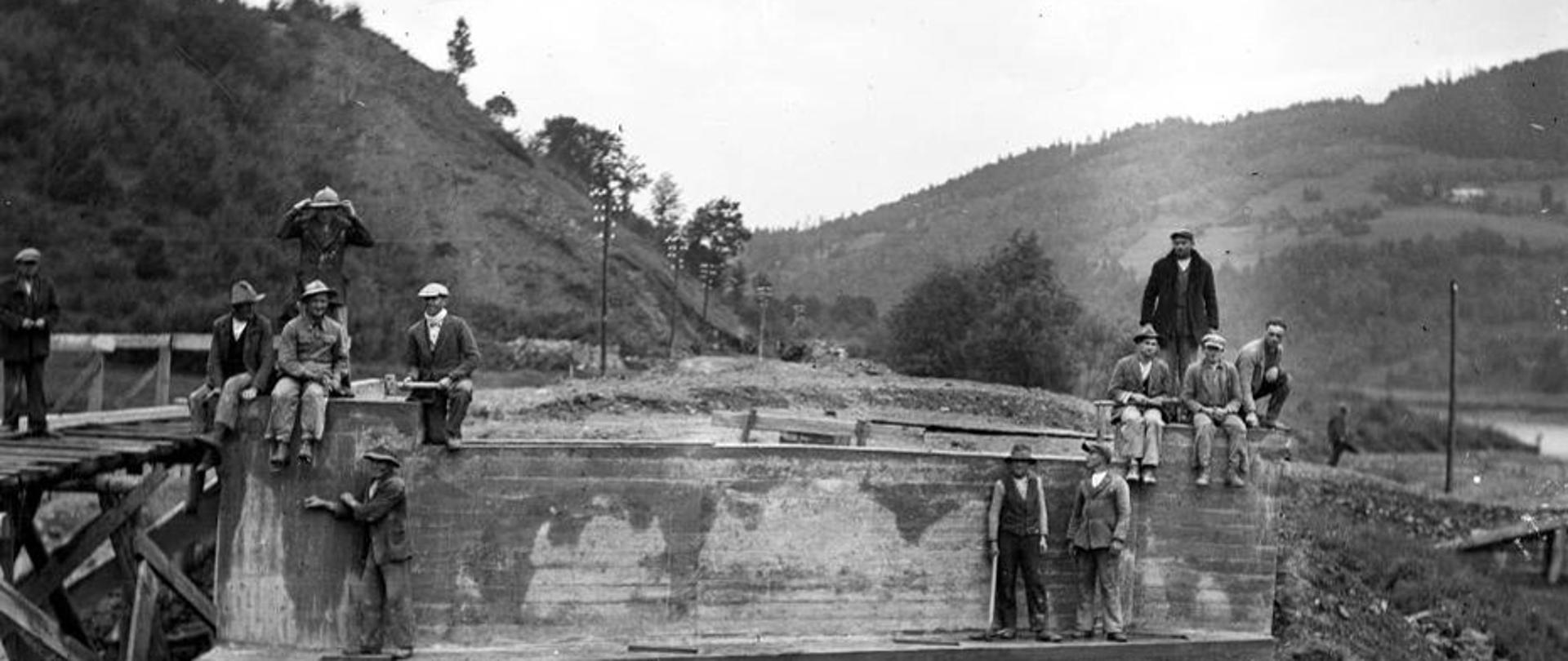 Czarno-białe zdjęcie na którym widoczni są robotnicy pracujący przy budowie Zakopianki. Panowie ubrani są w zwykłe ubrania robotnicze, na głowach mają kapelusze albo czapki. Sześcioro z nich siedzi, pozostali stoją.