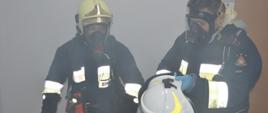 Szkolenie podstawowe strażaków-ratowników OSP. Wnętrze komendy powiatowej w Rawiczu. Dwóch strażaków - kursantów wykonuje ćwiczenie z użyciem sprzętu ochrony układu oddechowego. Strażak stojący z lewej strony jest w pełni przygotowany do realizacji zadania, natomiast jego kolega z prawej strony - zakłada hełm . Strażacy maja na sobie ubrania specjalne koloru ciemnego z elementami odblaskowymi. Na ich twarzach są maski oddechowe, a na plecach aparaty powietrzne. 
