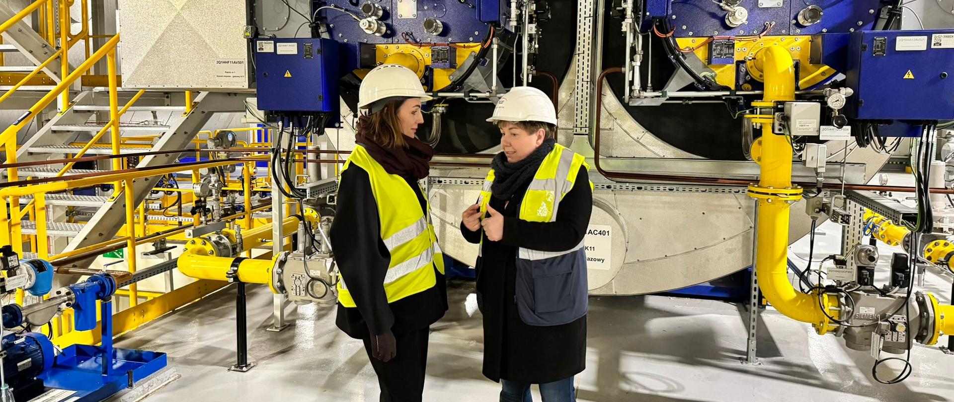 dwie kobiety stajo w pomieszczeniu elektrociepłowni, w tle rury i urządzenia elektroniczne 