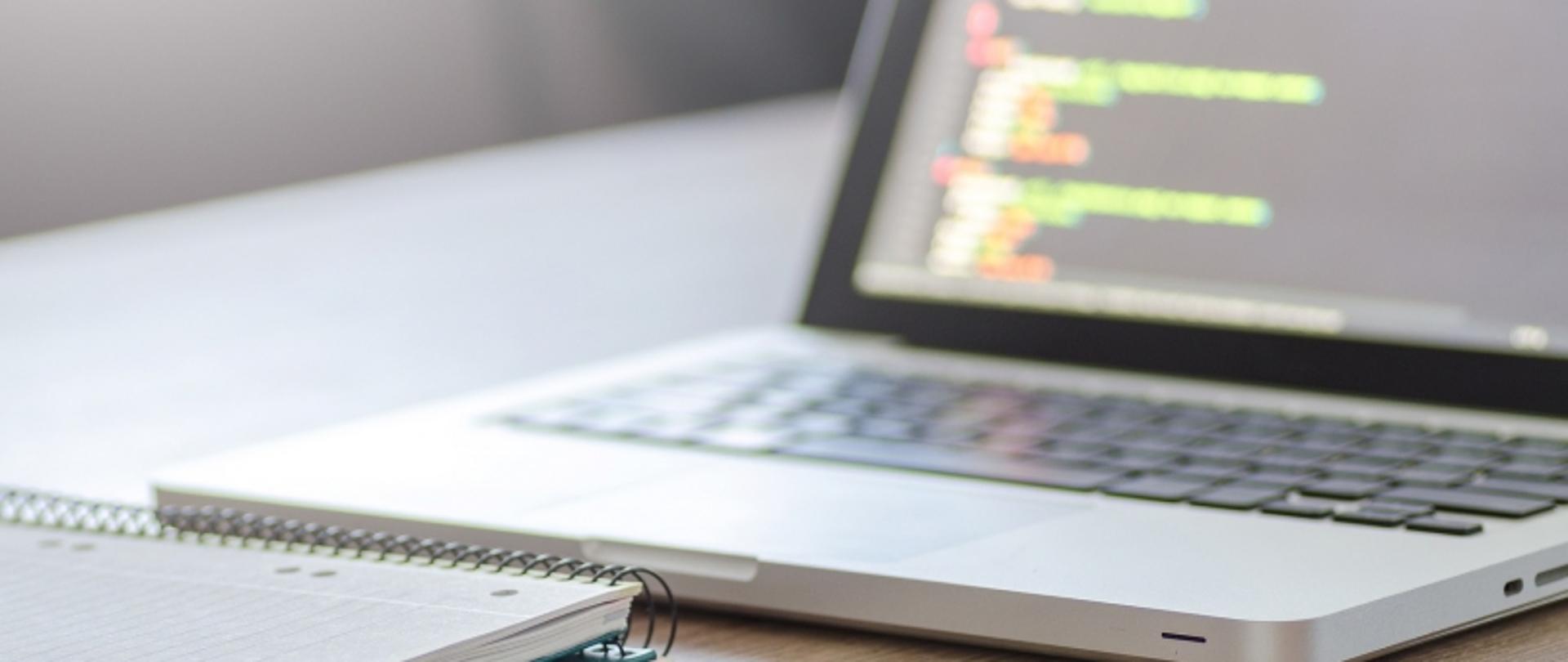 „Laptop z kodem na ekranie i notatnikiem na biurku.”