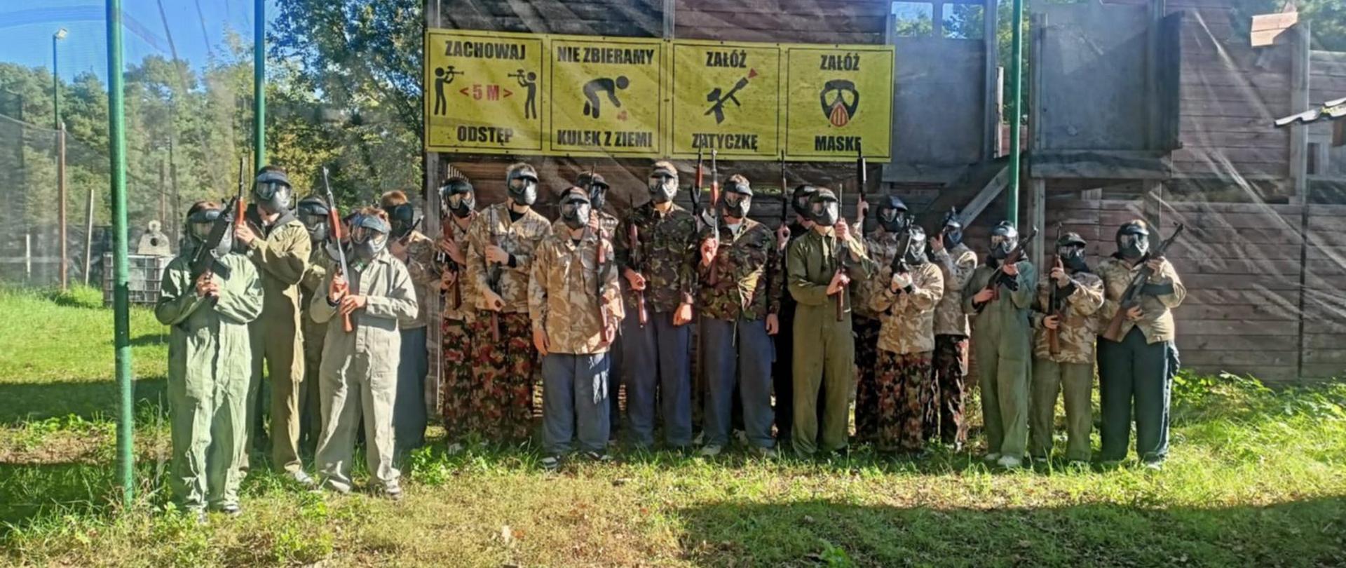 Na zdjęciu stoi duża grupa młodych ludzi- ponad 20- w pełnym umundurowaniu do gry w ASG. Wszyscy mają na sobie kombinezony w różnych odcieniach zieleni i brązów z elementami moro oraz maski ochronne zasłaniające twarz. Każdy trzyma w rękach replikę broni.