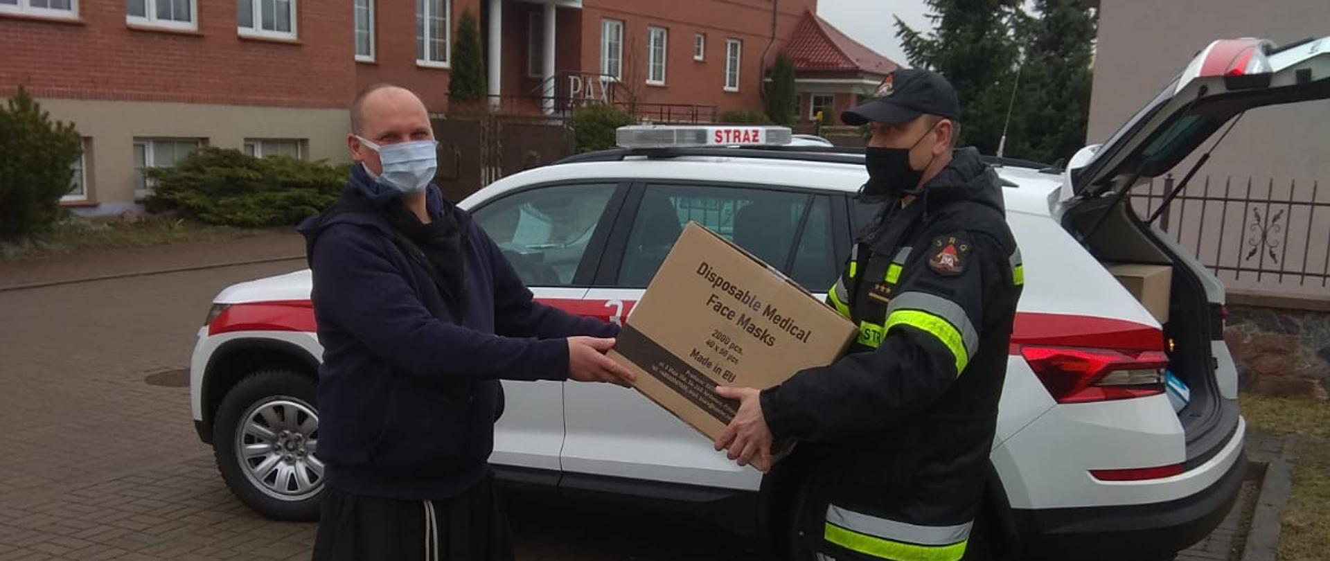 Zdjęcie obrazuje przekazanie księdzu przez strażaka maseczek ochronnych w tle widoczny samochód strażacki i część budynku.