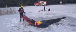 Na zdjęciu widoczne zamarznięty zalew w Dubiczach Cerkiewnych. w tle widoczny wóz strażacki, dwóch strażaków asekurujących sanie lodowe prowadzone przez strażaka. na saniach kolejny ratownik wciągający osobę poszkodowaną z przerębla.