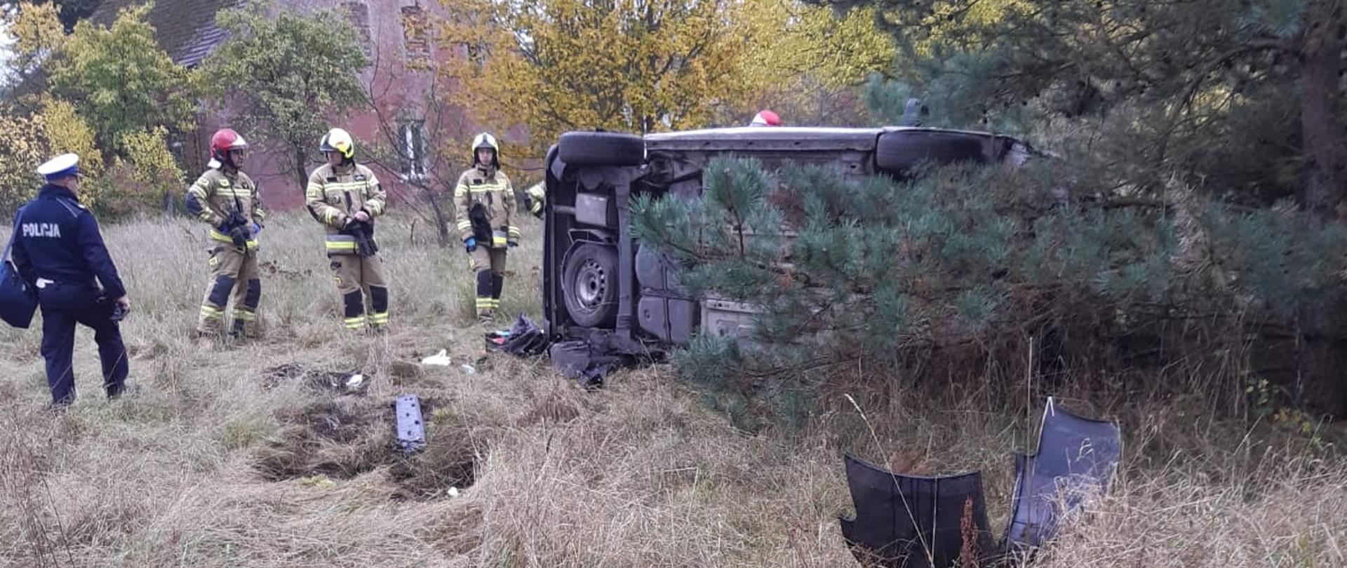 Na zdjęciu widać samochód leżący na boku. W tle strażacy w umundurowaniu specjalnym