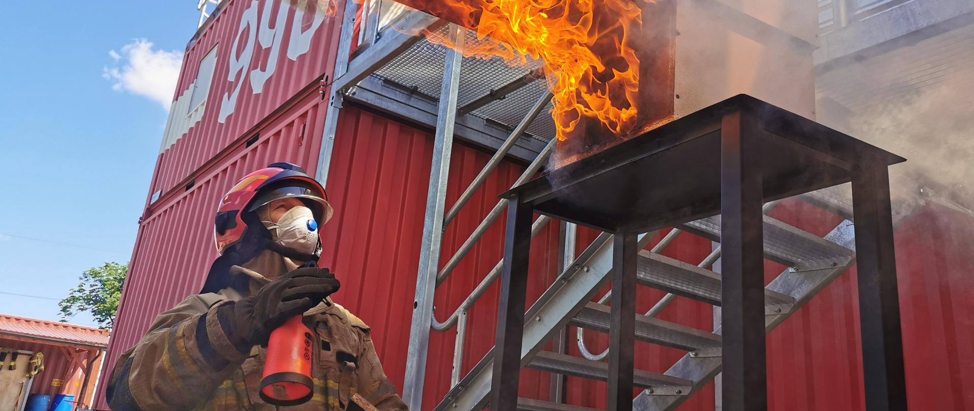 strażak w umundurowaniu specjalnym w hełmie i maseczce na twarxy niosący deskę , nad nim płomienie a w tle trenażer