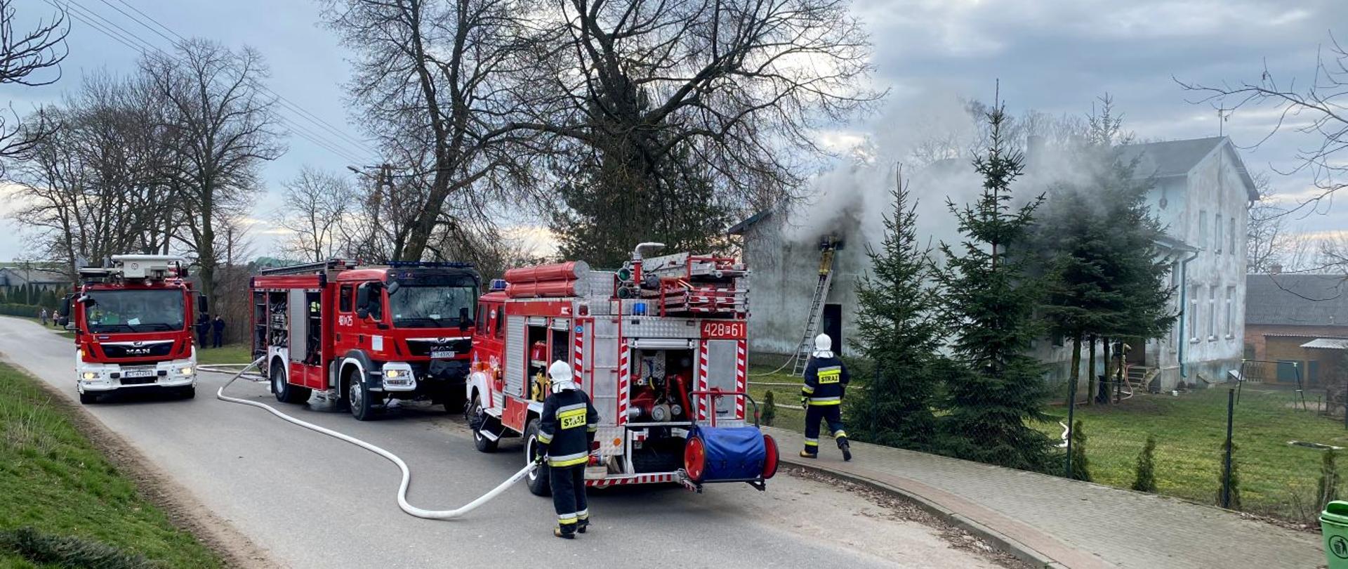 Na zdjęciu widać trzy samochody pożarnicze oraz rozwinięte linie gaśnicze. W tle widać zadymiony budynek w którym powstał pożar.