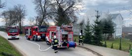 Na zdjęciu widać trzy samochody pożarnicze oraz rozwinięte linie gaśnicze. W tle widać zadymiony budynek w którym powstał pożar.