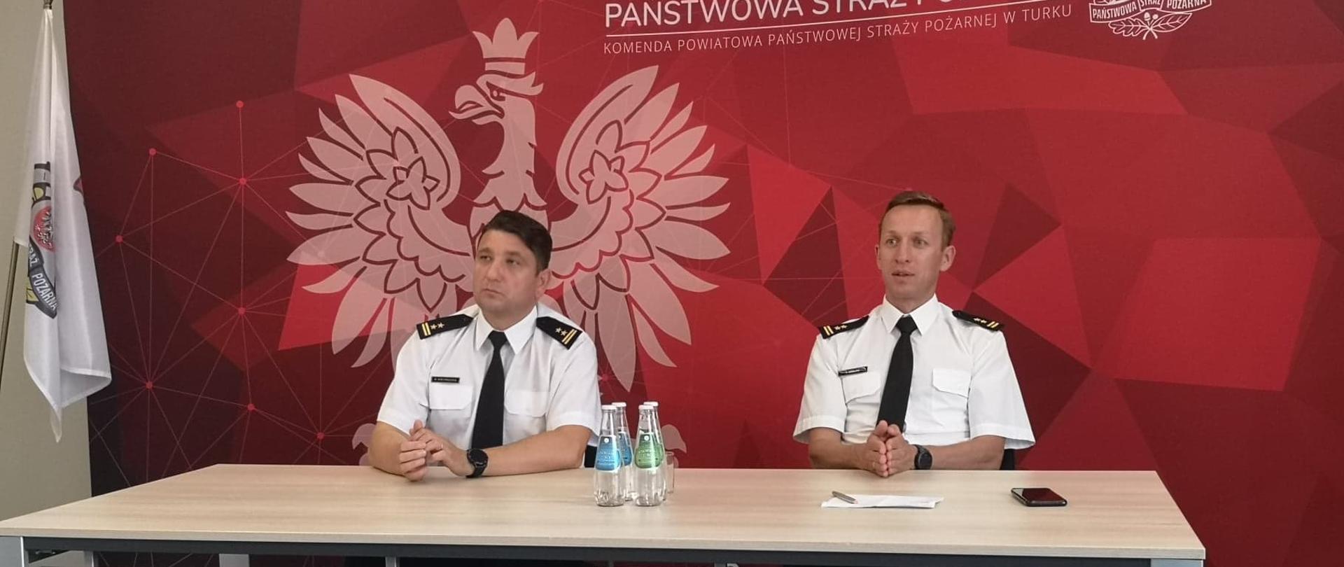 dwóch mężczyzn siedzi przed stołem w białych koszulach z czarnymi krawatami, za nimi czerwona ścianka z orłem