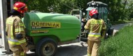 Zdjęcie przedstawia dwóch strażaków stojących przy ciągniku rolniczym z opryskiwaczem sadowniczy