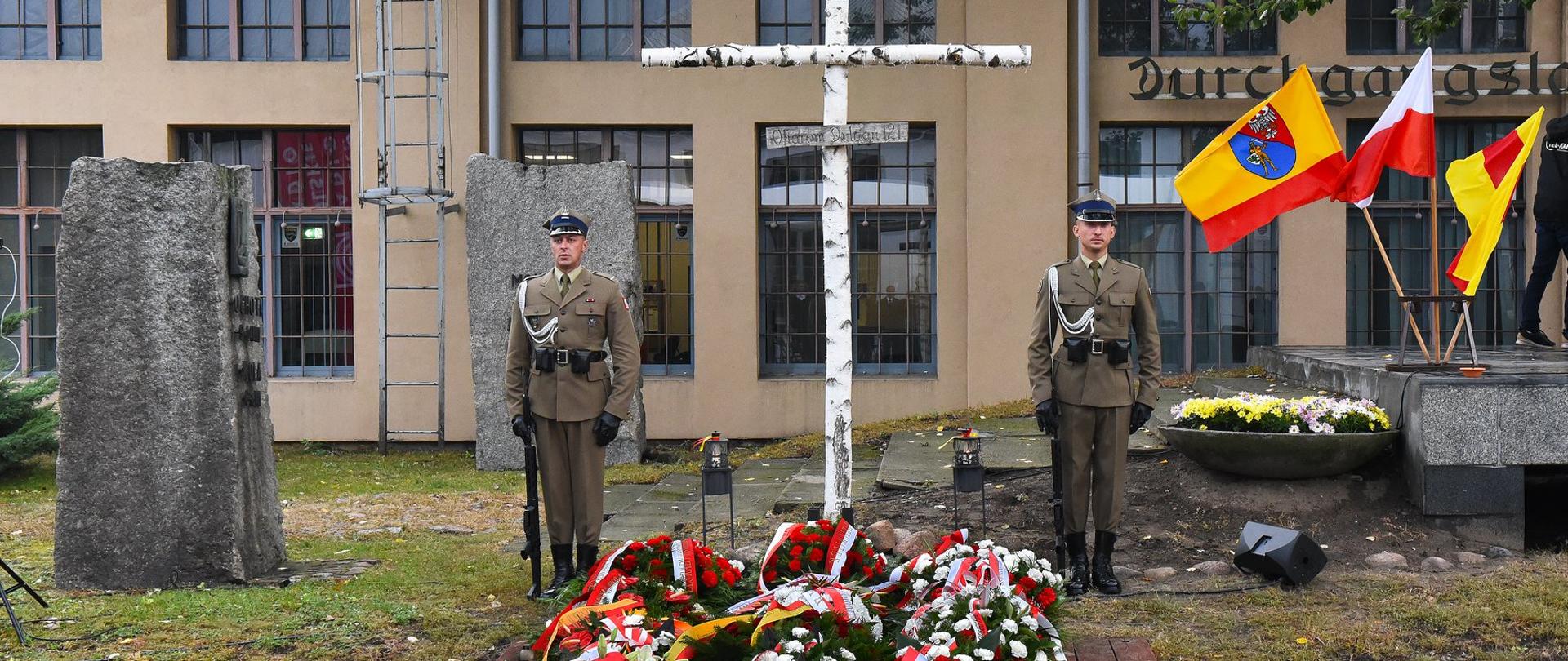 Zdjęcie przedstawia krzyż wykonany z brzozowych konarów, przed którym znajdują się biało-czerwone wieńce złożone przez delegacje. Przed krzyżem posterunek sprawują żołnierze Wojska Polskiego. W tle fragmenty pomnika „Tędy przeszła Warszawa” oraz budynek dawnych Zakładów Naprawczych Taboru Kolejowego