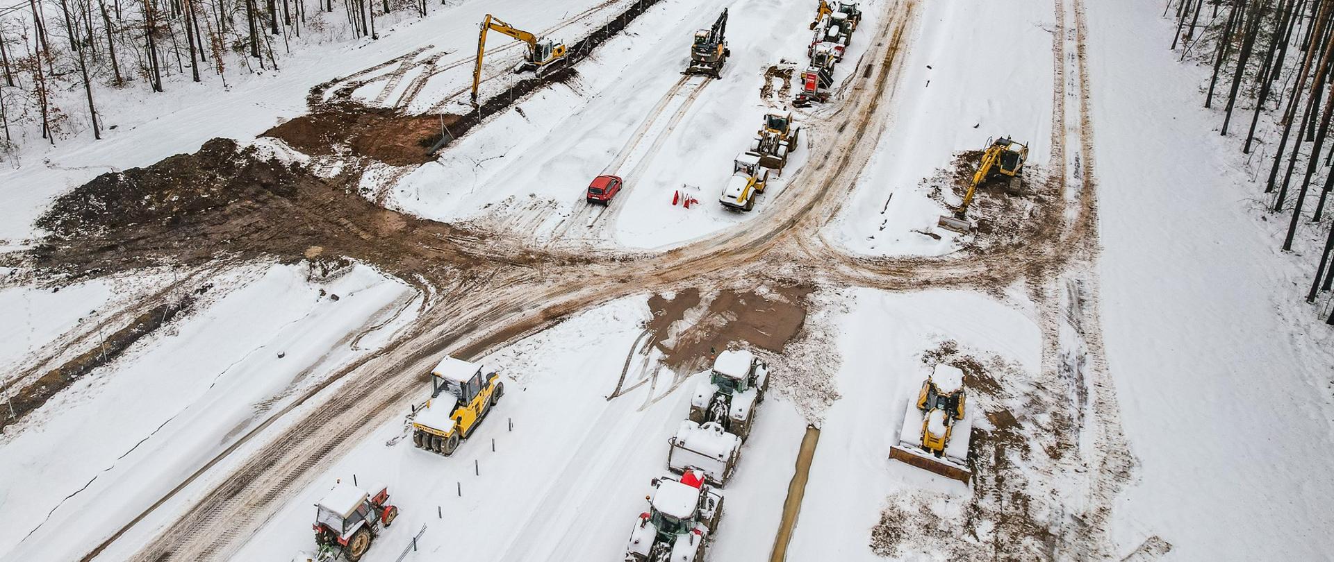 Na zdjęciu widzimy zaśnieżony plac budowy drogi, na którym pracują różne maszyny budowlane, w tym koparki, spycharki i walce. W tle znajduje się las, a teren pokryty jest śniegiem. Na pierwszym planie widać rząd pojazdów budowlanych oraz czerwoną osobówkę.