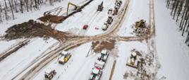 Na zdjęciu widzimy zaśnieżony plac budowy drogi, na którym pracują różne maszyny budowlane, w tym koparki, spycharki i walce. W tle znajduje się las, a teren pokryty jest śniegiem. Na pierwszym planie widać rząd pojazdów budowlanych oraz czerwoną osobówkę.