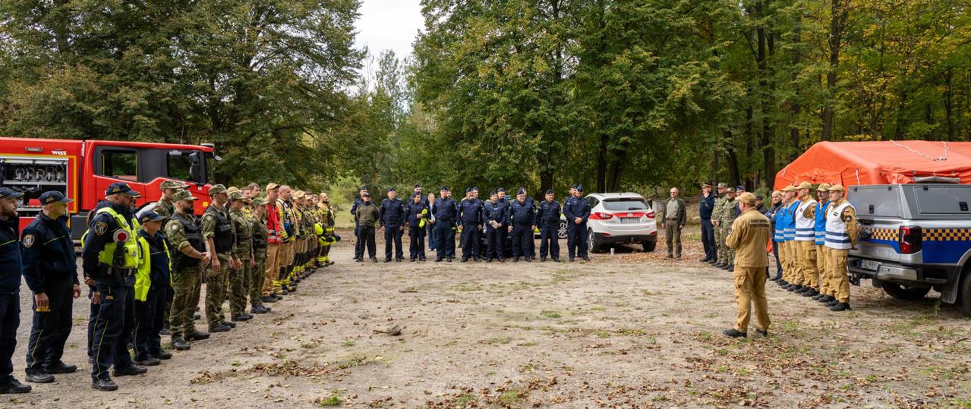 Komendant Miejski PSP przemawia podczas podsumowania ćwiczeń sprawdzających Miejski Plan Ratowniczy. W tle znajdują się przedstawiciele Straży Miejskiej, Państwowej Straży Pożarnej, Ochotniczej Straży Pożarnej, Policji oraz Straży Granicznej.