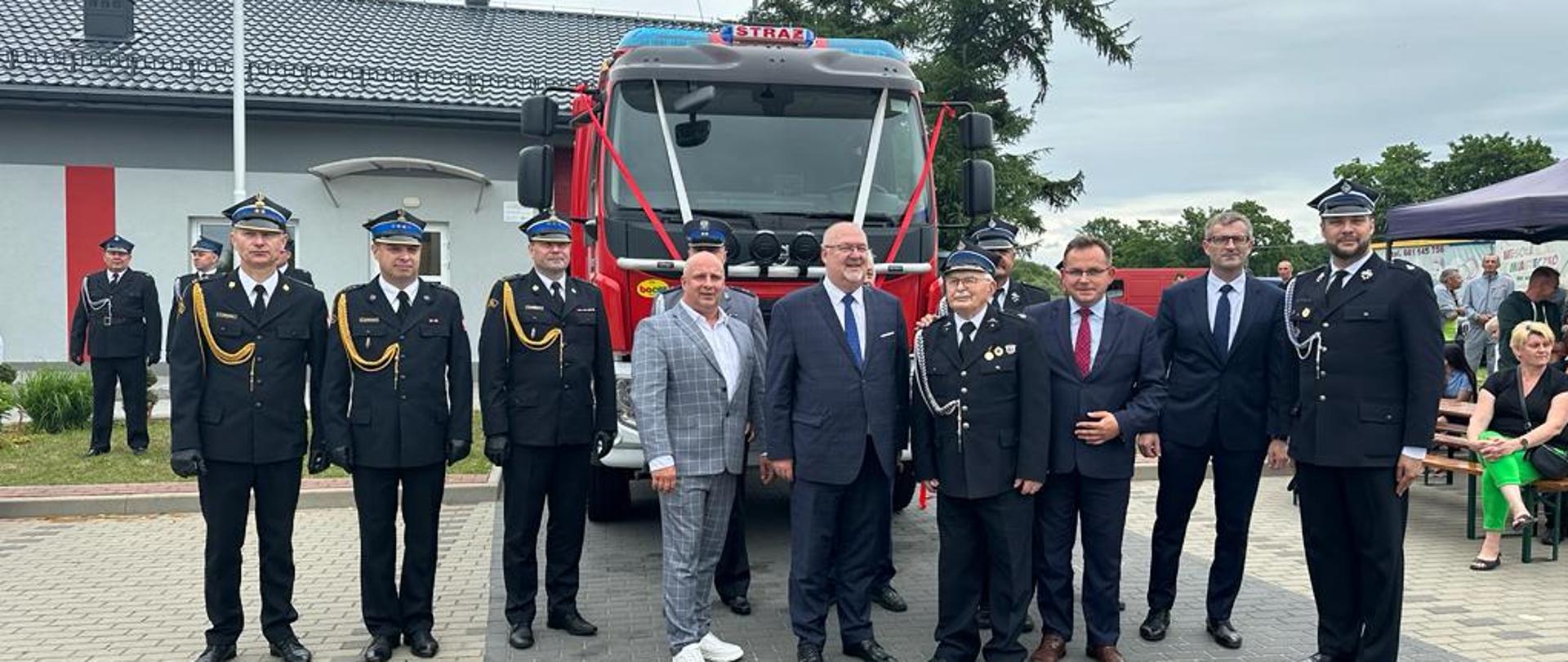 Przed wozem strażackim stoi 12 mężczyzn.