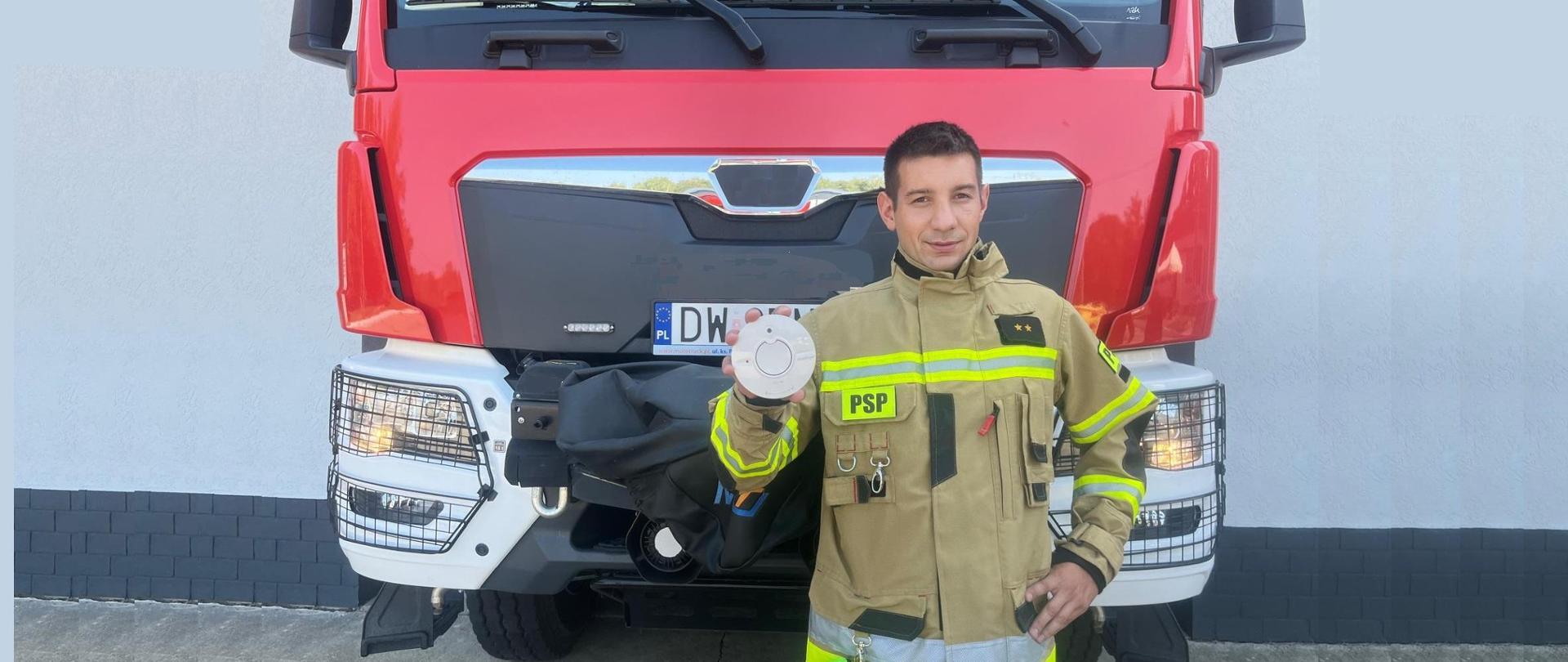 Strażak PSP z czujką w dłoni na tle samochodu gaśniczego