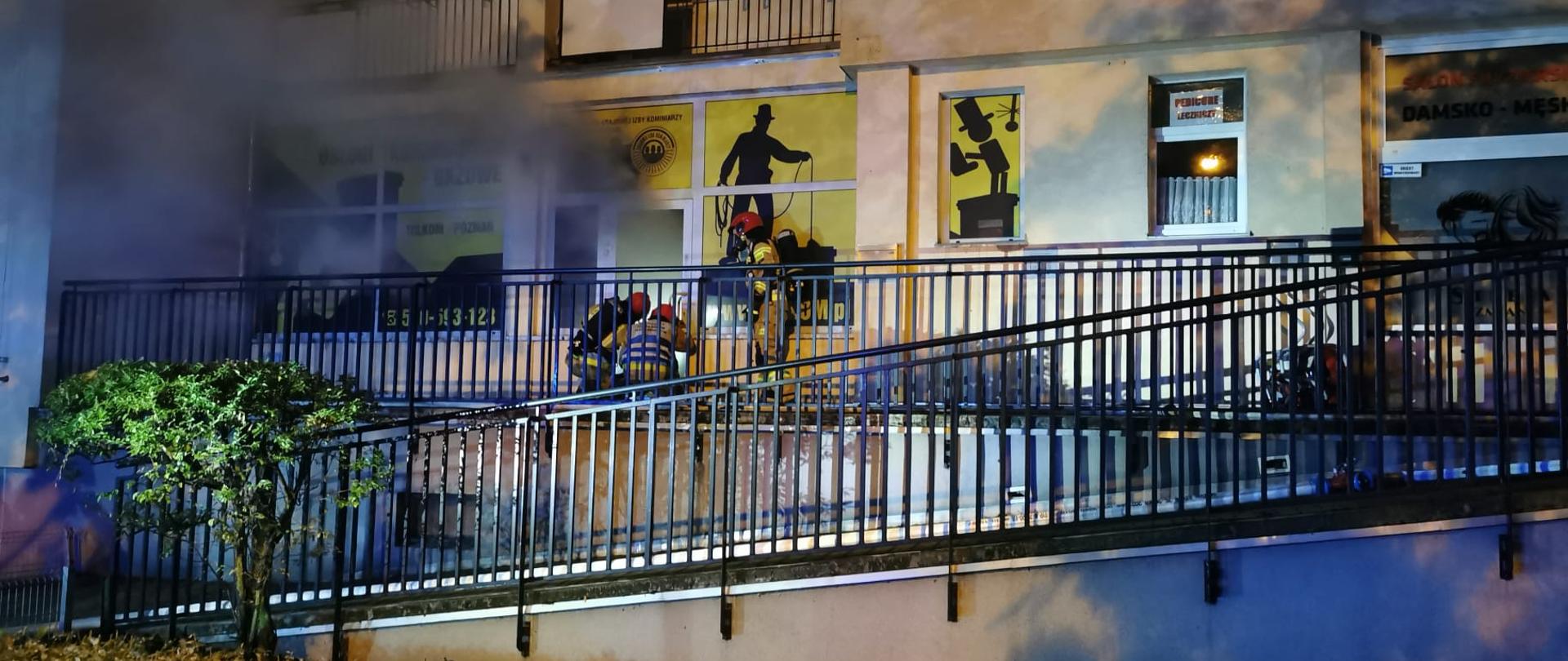 Zdjęcie przedstawia strażaków wchodzących do pomieszczenia usługowego w którym powstał pożar