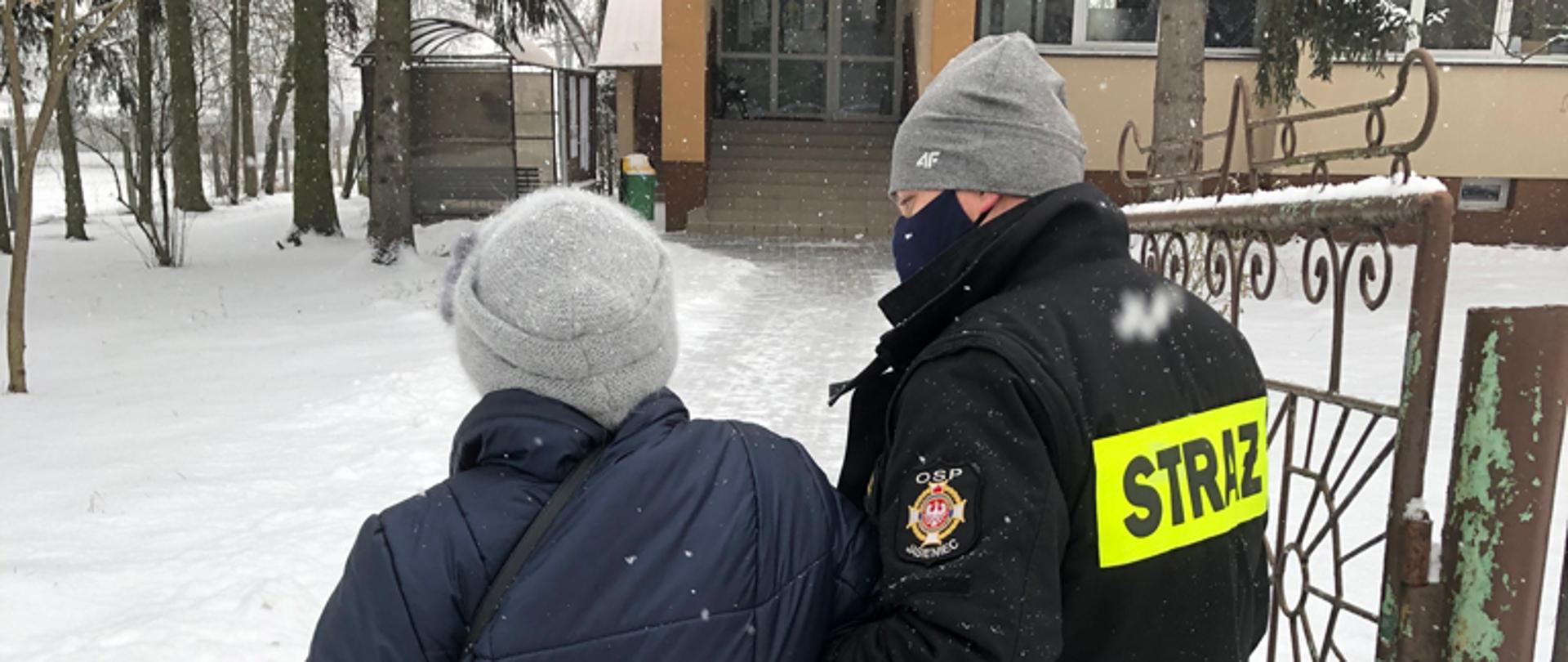 Strażak pomaga starszej osobie, prowadząc ją pod rękę do punktu szczepień.