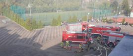 Na zdjęciu widać cztery samochody pożarnicze stojące na placu przy szkole podstawowej. W pobliżu samochodów kilku strażaków i druhów prowadzi działania związane z rozwinięciem linii gaśniczej i przygotowaniem sprzętu medycznego.