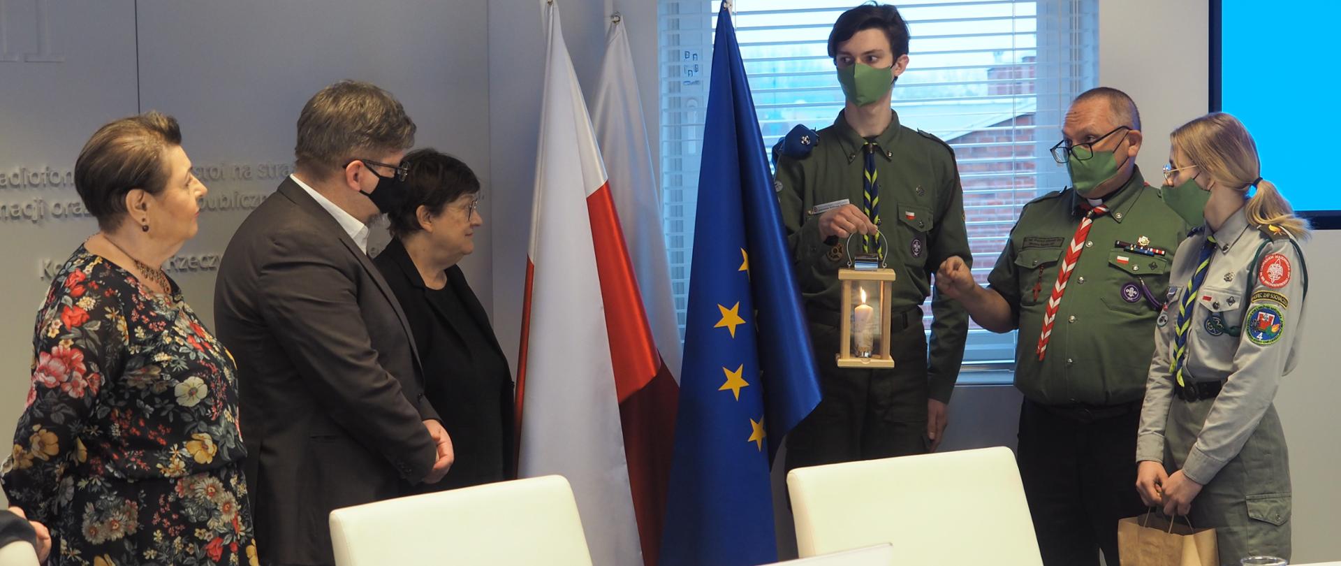 W sali posiedzeń KRRiT, po prawej stronie stoją harcerze i trzymają kaganek ze Światłem Pokoju. Po lewej stoją członkowie KRRiT. Po środku flagi Polski i Unii Europejskiej. 
