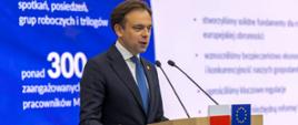 Minister Andrzej Domański stoi za mównicą z flagami Polski i Unii Europejskiej