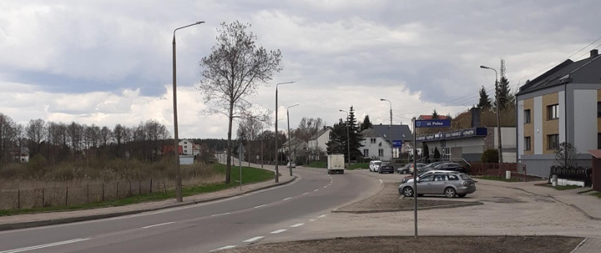 Łuk asfaltowej drogi, z prawej parkingi z autami i zabudowa 