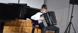 Na zdjęciu wykonanym w auli PSM widnieje uczeń grający na akordeonie, w tle fortepian. Kolorystyka zdjęcia jest biało-czarno-brązowa.