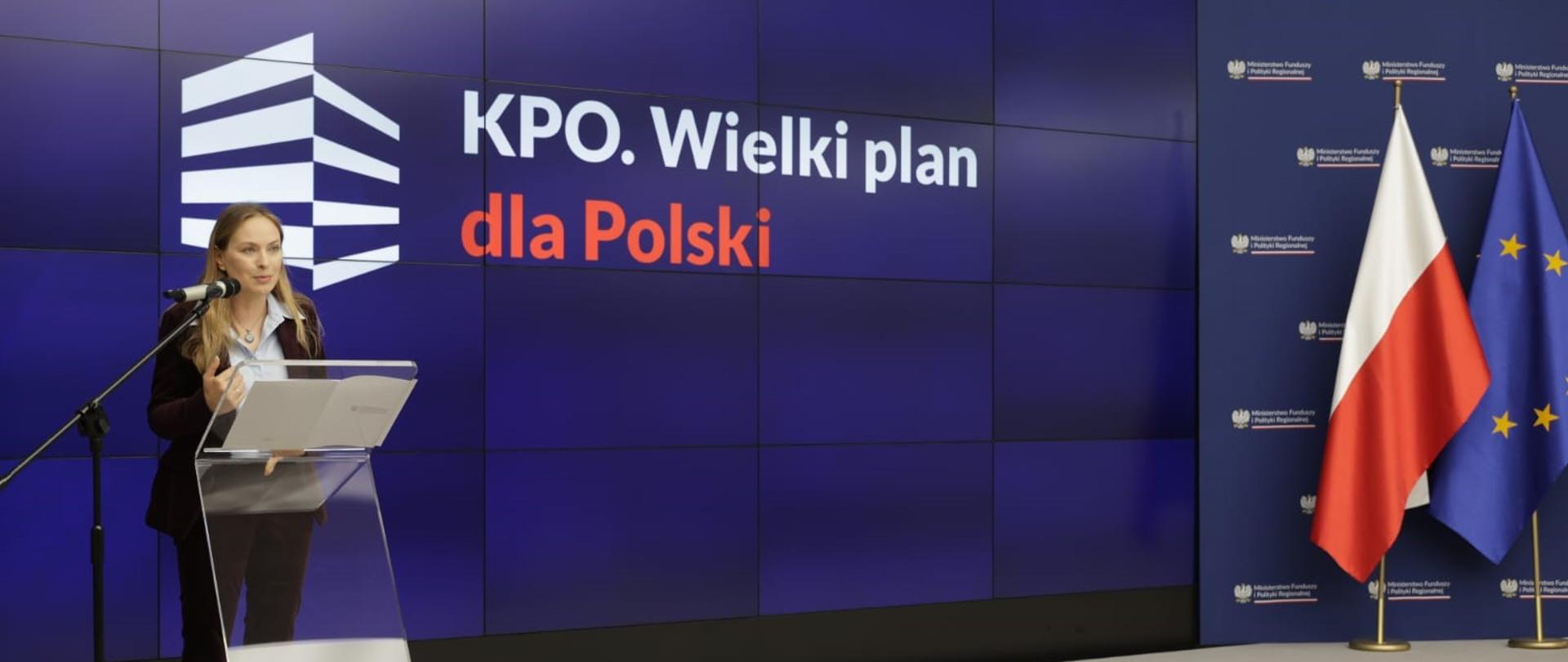 Minister Katarzyna Pełczyńska-Nałęcz stoi na podeście z lewej strony przy szklanej mównicy i mówi do mikrofonu. Z prawej strony sceny stoją flagi Polski i Unii Europejskiej. 
