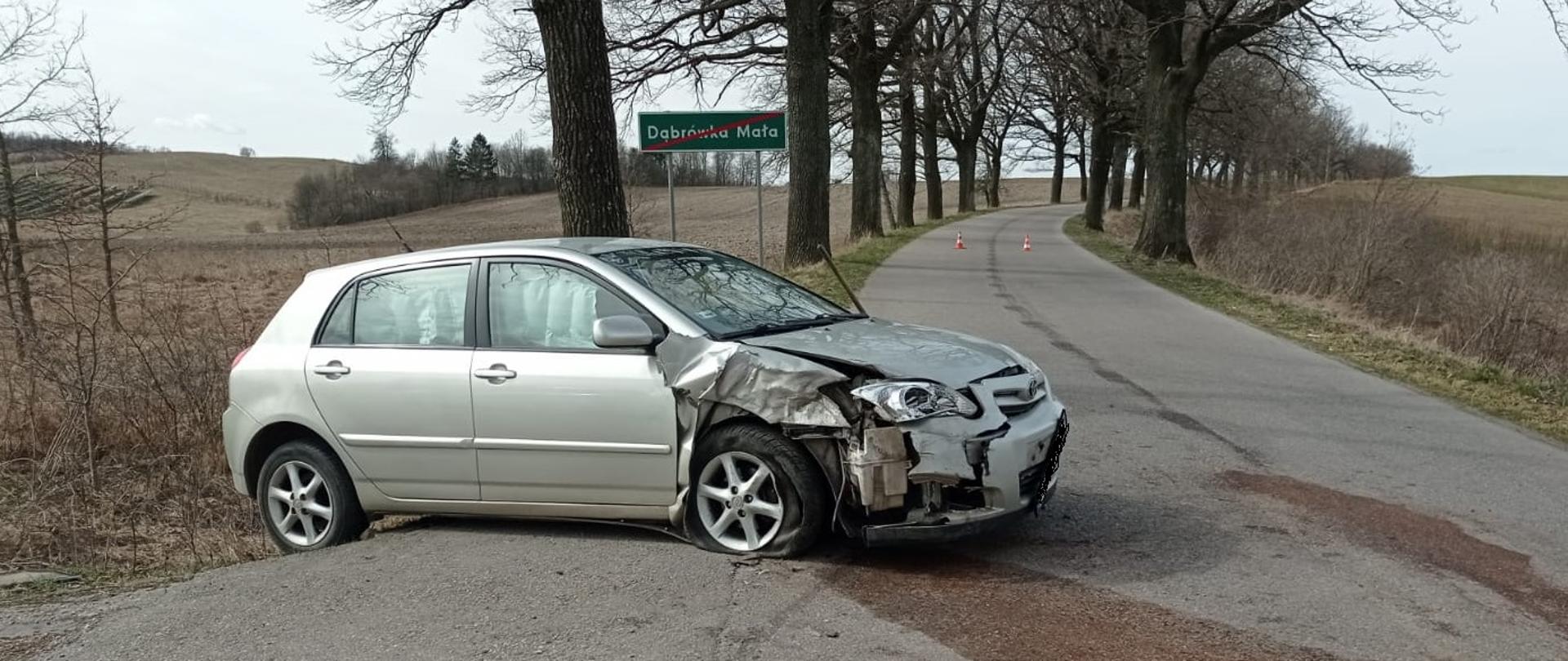 Srebrny z rozbitym przodem samochód osobowy stoi z boku drogi. Za nim znajduje się znak koniec miejscowości. 