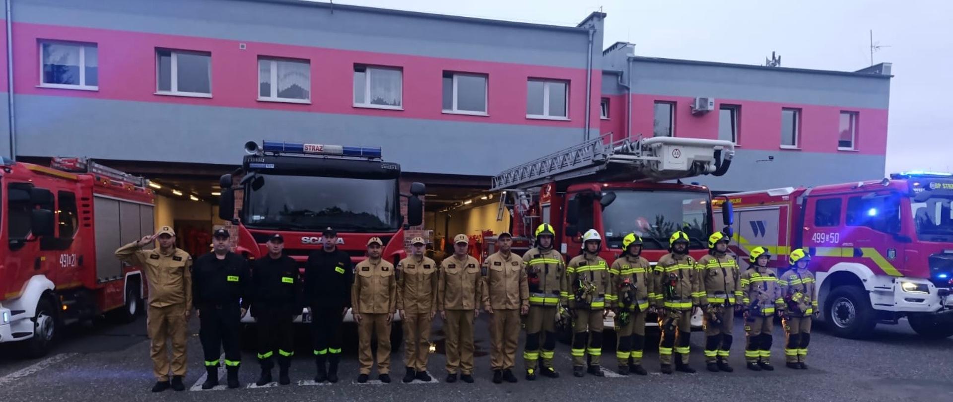Na zdjęciu znajdują się strażacy w mundurach służbowych i specjalnych, ustawieni na tle wozów strażackich i budynku komendy