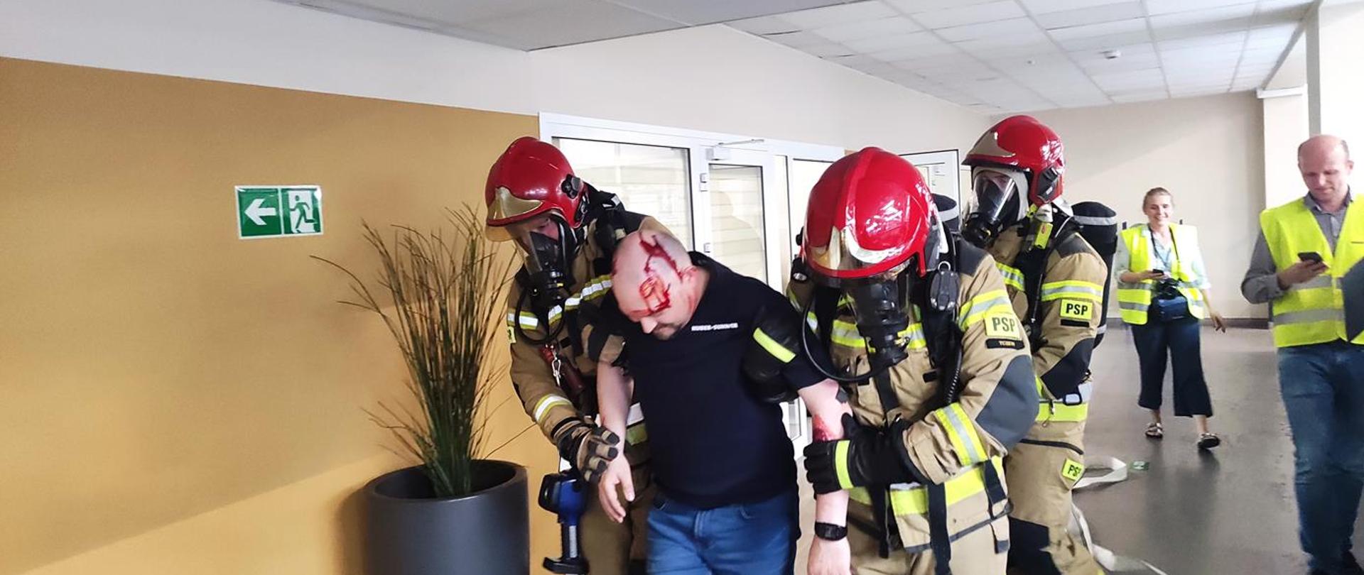 Korytarz firmy. Trzech strażaków w ubraniach specjalnych i maskach ochrony dróg oddechowych wyprowadza poszkodowanego pracownika, który ma ucharakteryzowany uraz głowy. Obok dwóch obserwatorów w kamizelkach.