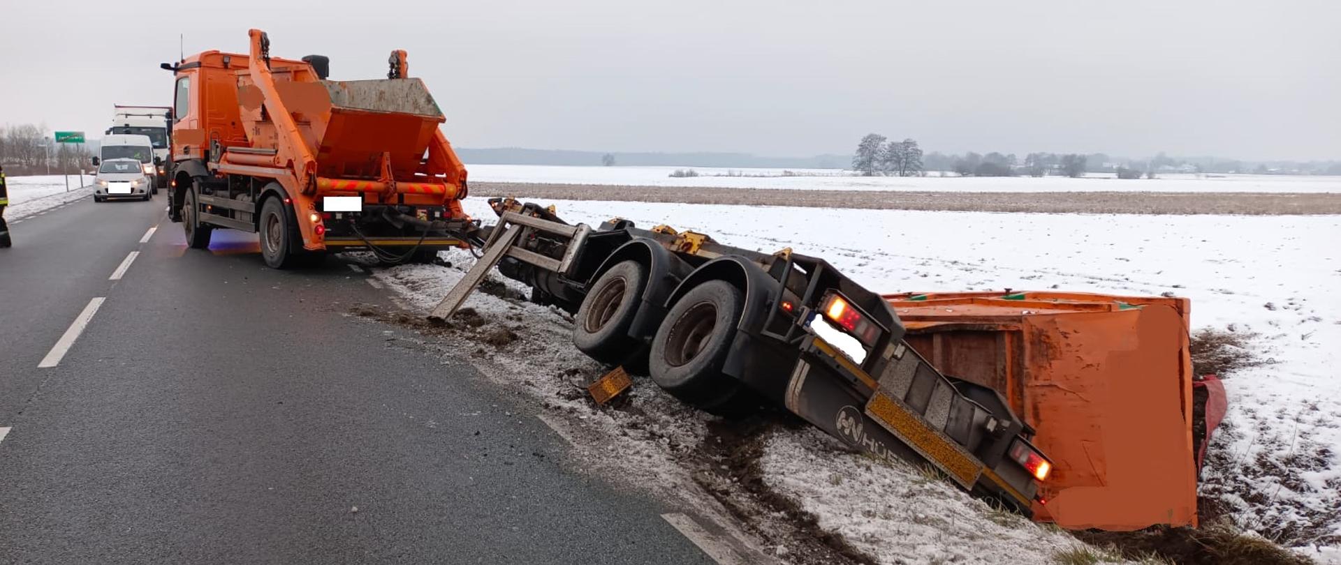 Widać samochód ciężarowy na drodze, który blokuje pas ruchu, przyczepa jest w rowie, kontener z przyczepy też w rowie, w około widać śnieg na polu