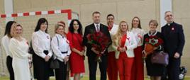 Zdjęcie przedstawia ministra Czarnka z bukietem czerwonych róż w towarzystwie kilkunastu kobiet ubranych w eleganckie i biało-czerwone stroje z kotylionami. Obok kobiet stoi także ksiądz (elegancko ubrany z przypiętym do garnituru kotylionem).