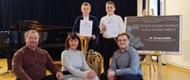 Na zdjęciu widzimy pięć osób pozujących na tle sceny z czarną kurtyną, fortepianem Yamaha oraz tablicą informacyjną o III Ogólnopolskim Konkursie Instrumentów Dętych, który odbył się w dniach 14–16 marca 2025 roku.
Na pierwszym planie, siedząc na stopniach sceny, znajdują się dwaj mężczyźni i kobieta. Mężczyzna po lewej stronie ma na sobie bordową koszulę i czarne spodnie, kobieta w środku nosi długą, szarą suknię, a mężczyzna po prawej stronie ubrany jest w niebieską koszulę i ciemne spodnie.
Za nimi stoją dwaj młodzi chłopcy, każdy trzymający dyplom i uśmiechający się do kamery. Przed nimi znajduje się tuba – instrument dęty blaszany. Cała grupa wygląda na zadowoloną, a otoczenie sugeruje uroczysty charakter wydarzenia.