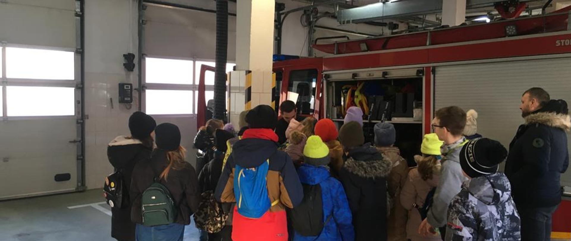 Zdjęcie przedstawia grupę dzieci wraz z opiekunem podczas wizyty w PSP. W tle samochód pożarniczy, otwarte skrytki i drzwi.