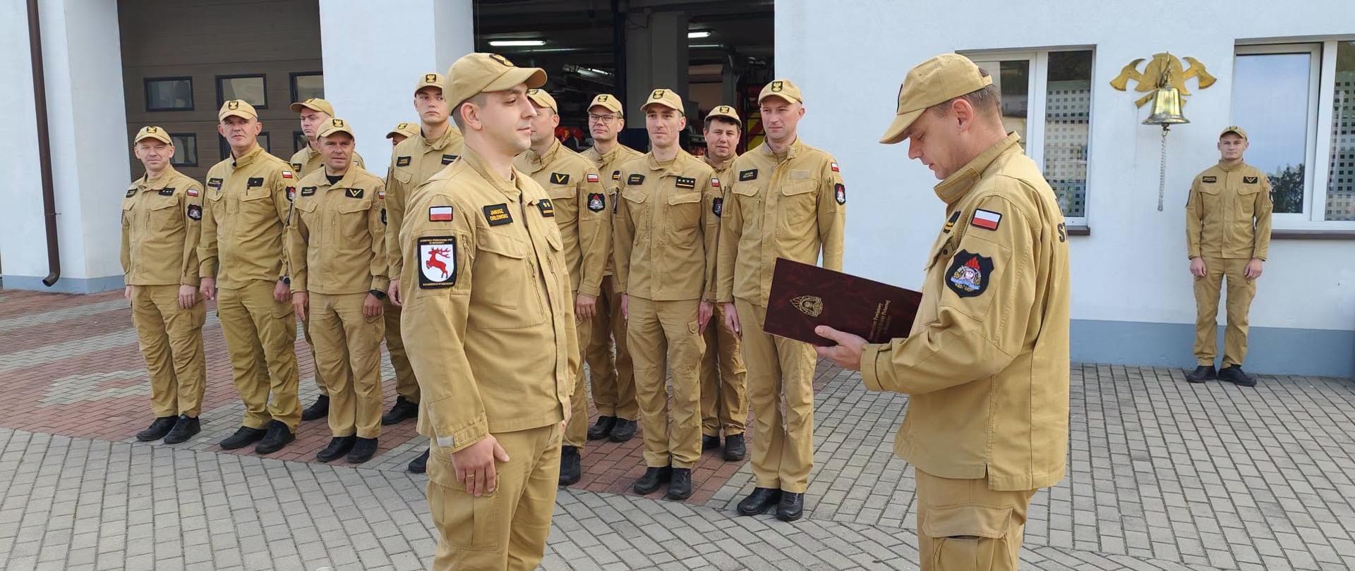 Zdjęcie przedstawia Komendanta Powiatowego PSP w Brodnicy mł. bryg. Krzysztofa Natuckiego wręczającego mianowanie na wyższe stanowisko służbowe mł. kpt. Januszowi Orłowskiemu. W tle w dwuszeregu stoją funkcjonariusze Komendy. Wszyscy ubrani w umundurowanie służbowe Straży Pożarnej w kolorze miodowym.