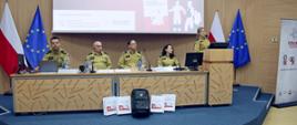 Od lewej trzech funkcjonariuszy PSP oraz funkcjonariuszka PSP siedzący za stołem prezydialnym w auli wykładowej podczas konferencji, po prawej mównica z której prelekcję wygłasza funkcjonariuszka PSP. W tle ekran multimedialny z wyświetlonym slajdem prezentacji