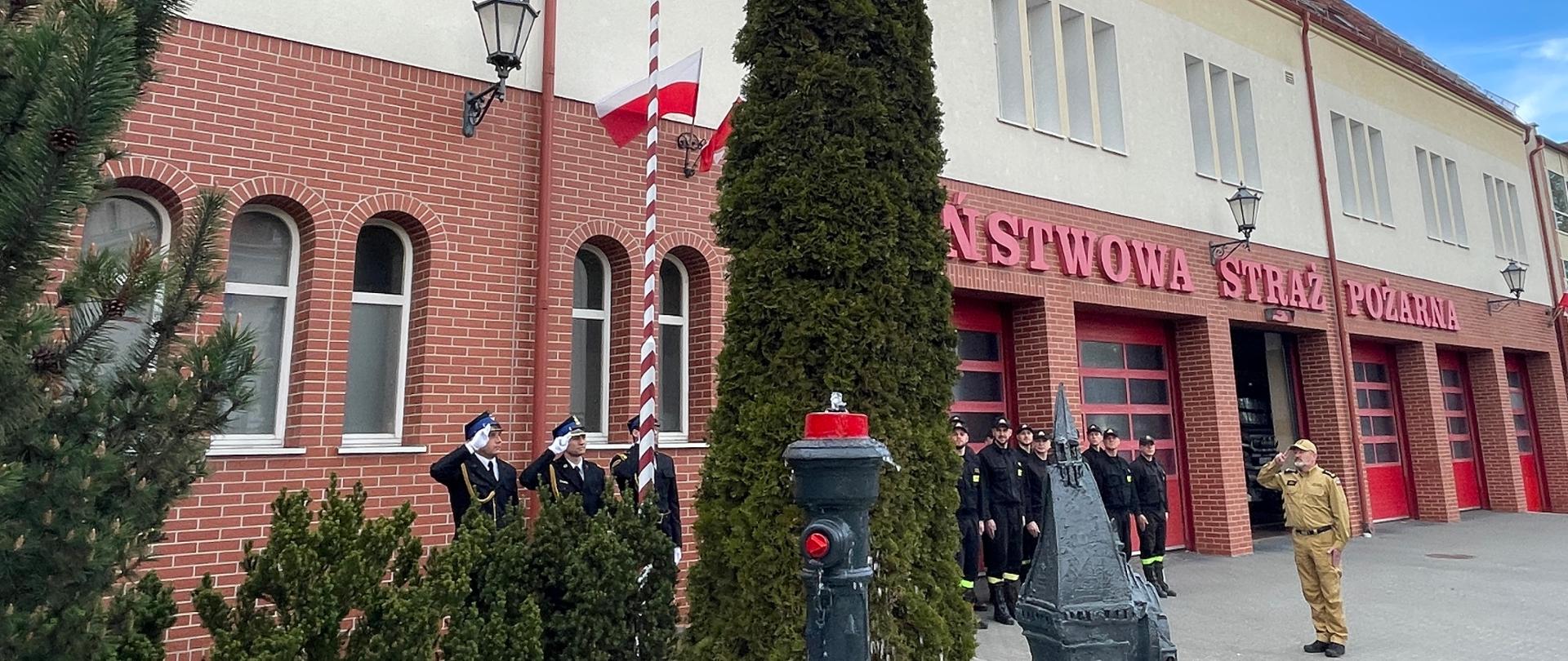 strażacy stojący w dwuszeregu przed garażami a przed nimi komendant. po lewej poczet flagowy wciąga flagę na maszt
