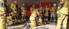 Strażacy z Państwowej Straży Pożarnej podczas ćwiczeń z zakresu RIT (Rapid Intervention Team). Dziesięciu strażaków w piaskowych mundurach bojowych otacza leżącego na płycie instruktora, który demonstruje techniki stabilizacji lub ewakuacji. W tle widać czerwone pojazdy bojowe. Na pierwszym planie na podłodze leżą hełmy i zestawy aparatów oddechowych z noszakami.