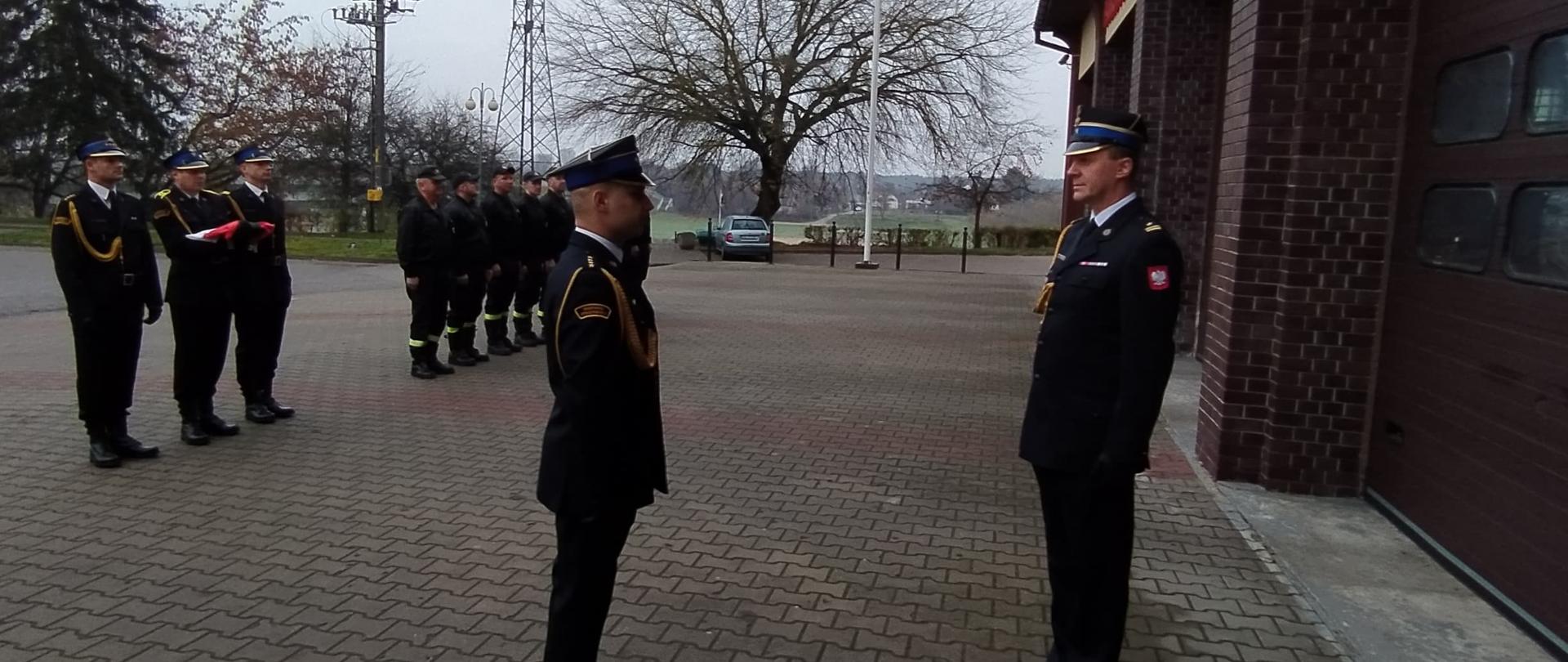 Dowódca składa meldunek Komendantowi. W tle szereg strażaków i poczet flagowy.