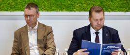 Minister Czarnek i mężczyzna w jasnobrązowej marynarce siedzą za białym stołem, minister czyta niebieską książkę z napisem Biznes i zarządzanie.