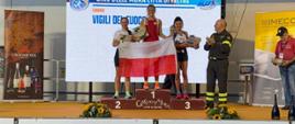Podium zawodów, na którego stopniach stoją 3 kobiety. Dwie trzymają biało – czerwoną flagę.