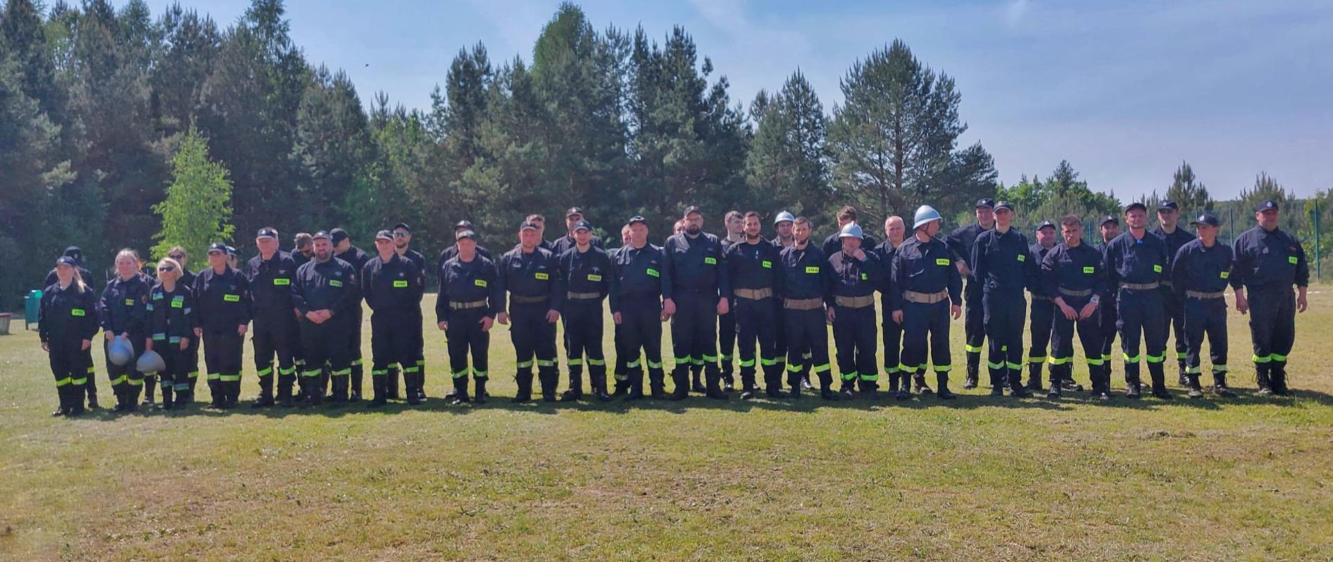 Na zielonej trawie w dwuszeregu stoi kilkudziesięciu strażaków.