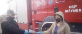 Zdjęcie przedstawia strażaka OSP, który wręcza kobiecie ulotkę dotyczącą szczepień przeciwko COVID-19. Zdjęcie wykonane jest na tle pojazdu strażackiego. Widać kabinę pojazdu, na której jest napis OSP Giżynek oraz znajdują się numery operacyjne 519c25. 