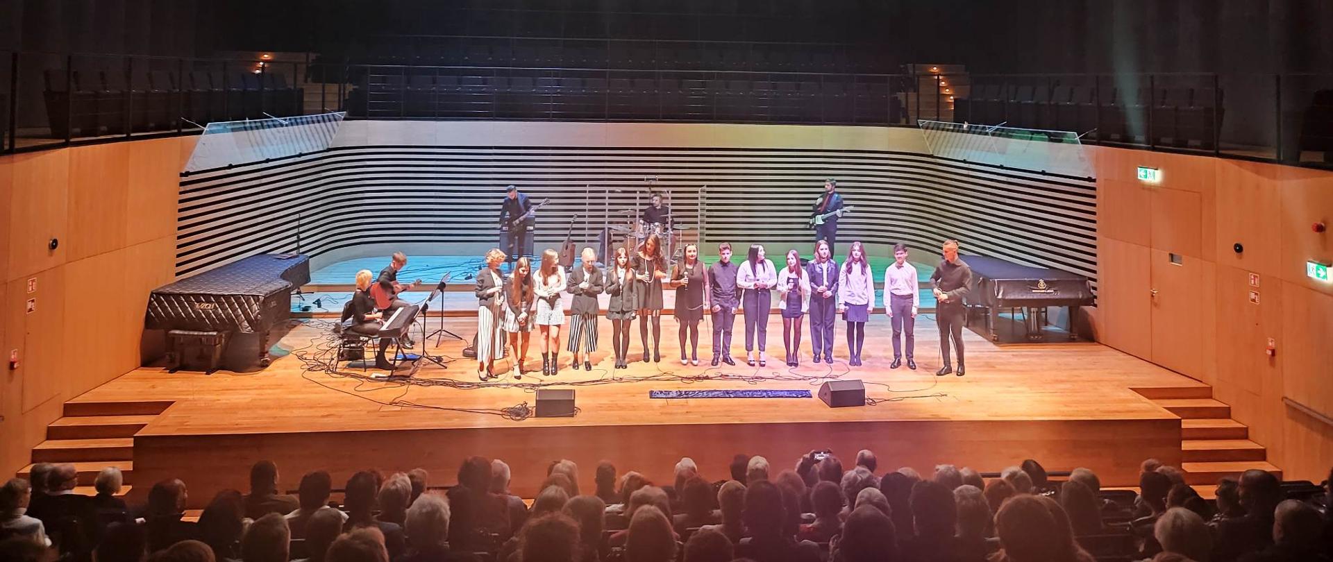 14 osób stoi na scenie sali koncertowej, po lewej kobieta siedzi przy pianinie cyfrowym, mężczyzna gra na gitarze, z tyłu sceny perkusista gra na perkusji, dwóch mężczyzn gra na gitarach elektrycznych