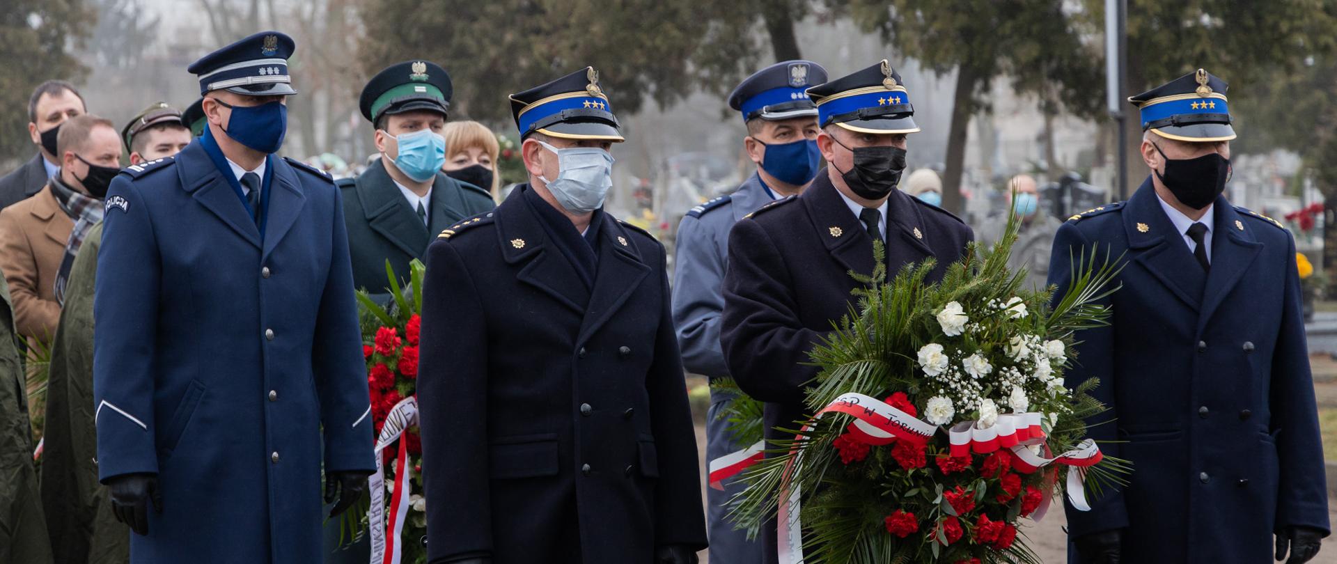 Delegacja PSP w płaszczach umundurowania wyjściowego, 3 osoby. Środkowa osoba trzyma wiązankę biało-czerwonych kwiatów. Wszyscy w maseczkach. W tle delegacje innych służb i instytucji
